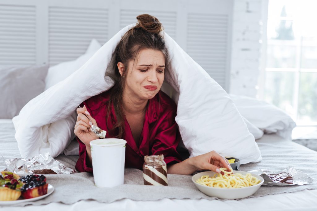 Crying woman lying in her bed surrounding by snacks